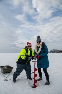 Two scientists pause from drilling into the ice on a frozen body of water to smile at the camera