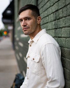 Aram Mrjoian standing against a green brick wall outdoors, wearing a light-colored button-up shirt and looking toward the camera with a neutral expression Aram Mrjoian standing against a green brick wall outdoors, wearing a light-colored button-up shirt and looking toward the camera with a neutral expression