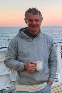 A man in a grey sweatshirt smiles on the bow of a boat with a white mug in his hands A man in a grey sweatshirt smiles on the bow of a boat with a white mug in his hands