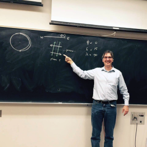 A man in glasses stands in front of a chalkboard with writing on it