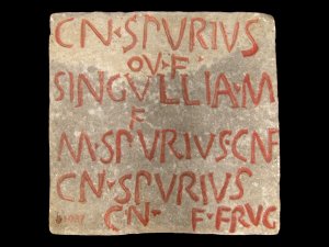 Square piece of coarse-grain white marble with lettering that is painted with red pigment. The Latin inscription reads, “CN SPVRIVS / OV F / SINGVLLIA M / F / M SPVRIVS CNF / CN SPVRIVS / CN F FRVG.”