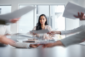 Woman sitting at a conference table looking stressed, with papers and conversations blurred, signaling motion.