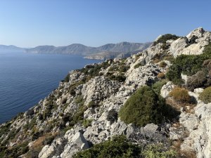 A rocky hillside with low-lying green/brown shrubs, sloping to a deep blue sea and a chain of islands/coastline in the background.