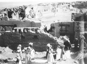 The archaeological site of Karanis showing partial brick walls of structures. Egyptian men and women, many of whom carry baskets, walk in front of and within the ruins. The archaeological site of Karanis showing partial brick walls of structures. Egyptian men and women, many of whom carry baskets, walk in front of and within the ruins.