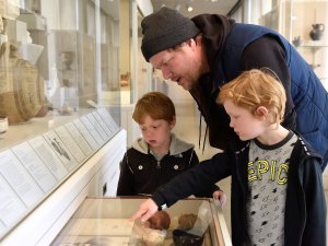 An adult and two children in the Kelsey Museum’s Greek gallery. They look at a drawer full of Greek pottery vessels and sherds pulled out from beneath the main display case. An adult and two children in the Kelsey Museum’s Greek gallery. They look at a drawer full of Greek pottery vessels and sherds pulled out from beneath the main display case.