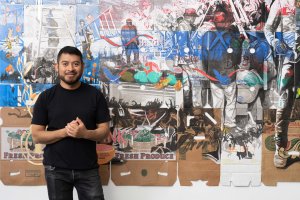 Narsiso Martinez stands in front of a colorful mural made from repurposed cardboard boxes. The art depicts scenes of workers and produce. Narsiso Martinez stands in front of a colorful mural made from repurposed cardboard boxes. The art depicts scenes of workers and produce.