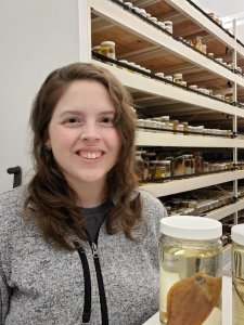 Photograph of Emily Troyer wearing a grey zip-up pullover, with scientific specimens in jars in the background.