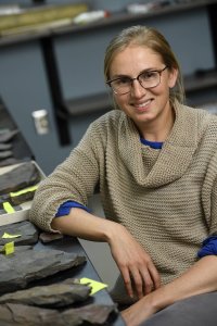 A woman in glasses smiles at the camera while sitting in a lab space with rocks on the table in front of her