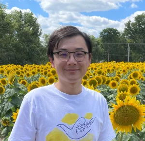 A person in glasses smiles in front of a field of sunflowers