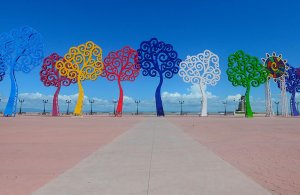 Vibrant metal tree sculptures in red, yellow, blue, and green line a pink walkway under a clear blue sky. Vibrant metal tree sculptures in red, yellow, blue, and green line a pink walkway under a clear blue sky.