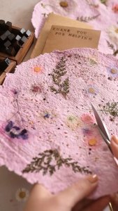 this image depicts hands cutting pieces of seed paper stacked together. the paper appears to be handmade and contains seeds and bits of flowers.