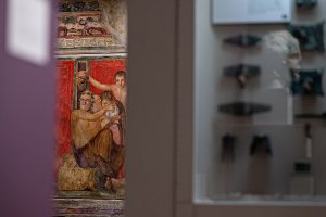 Partial view into the Kelsey Museum’s Villa of the Mysteries room, featuring brightly painted watercolor reconstructions of the Pompeiian frescoes. The visible panel shows a satyr holding up a theater mask, with Silenus offering another satyr a bowl to drink from.