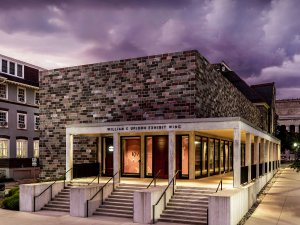 The Kelsey Museum’s William E. Upjohn Exhibit Wing against a cloudy purple sky in the evening. The brick building features a flat roof, a triple staircase, and a long porch.