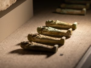 A group of four small, greenish, bearded ancient Egyptian ushabtis lying down in a row in a Kelsey Museum display case.