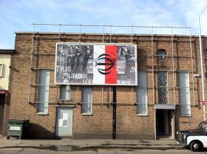 A photograph featuring figures from the Solidarity movement and a Polish white and red flag hanging on a brick wall of a building.