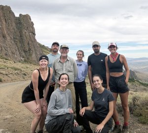 A group of eight people are posing together on a dirt path in a rugged, mountainous landscape. They are dressed in casual hiking clothes, with some wearing hats or bandanas, indicating they are on an outdoor adventure. Behind them, steep rocky cliffs rise to the left, and the scene opens to a vast valley and distant hills under a cloudy sky. The ground is dry and covered with sparse grasses and shrubs.