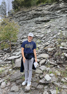A woman in a hat and blue Michigan EARTH t-shirt stands in front of a rock wall outside