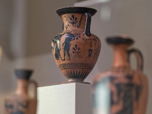 Museum case of black-figure Greek pottery. Prominently shown is a two-handled pot with two human painted figures on it.