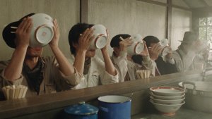 A movie still showing a line of Japanese people slurping ramen in a ramen shop