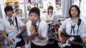 Four highschool girls in a rock band are mid-performance in a school, classroom