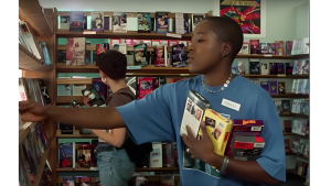 A person wearing a blue shirt and name tag browses a video rental store, holding several VHS tapes. Shelves filled with colorful movies surround them.