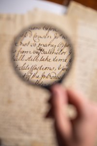 A hand holds a magnifying glass over an old handwritten manuscript, enlarging the cursive text on aged paper while the surrounding page remains softly out of focus.