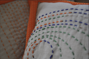A close-up of a quilted fabric with a bright orange boarder and an embroidered quilt square. The quilt square has a orange, blue, and green running stitches in concentric circles.