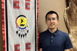 Photo of Wesley Leonard, Native American man with short dark hair and wire glasses, standing by tribal banner.