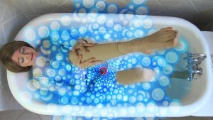 A woman reclines in a bathtub, with blue bubbles rising from the water