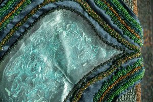Close-up detail of a textile and mixed-media artwork featuring layered fabrics and beadwork. A central curved form is covered in a pale blue-green, semi-translucent material with textured, marbled surface patterns. Surrounding it are bands of dark blue fabric densely embroidered with rows of green and gold beads that follow curved, wave-like lines. The beads vary in size and create a raised, shimmering surface. The composition emphasizes texture, stitching, and color contrast between the smooth central surface and the intricate beaded borders.