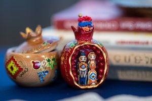 Close-up photograph of two small decorative Armenian objects displayed on a table. On the right is a red, pomegranate-shaped ornament opened to reveal painted figurines of a man and a woman wearing traditional Armenian clothing, framed by small red bead details. On the left is a light wooden pomegranate-shaped container with a removable lid, decorated with hand-painted Armenian motifs in red, green, blue, and yellow. Blurred books are visible in the background, emphasizing the objects as cultural artifacts associated with memory and tradition.