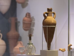 An amphora, a glass flask, and cups on display at the Kelsey Museum. More transport amphorae are visible in the background, mounted on a wall.