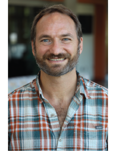 Broxton Bird wearing a blue, orange, and white plaid button-up shirt stands indoors with a blurred background featuring soft lighting and indistinct objects.