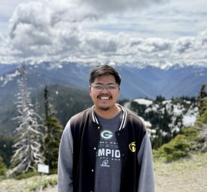 A man in glasses and a black coat stands in front of a mountain range