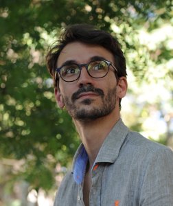 Man in glasses and a grey shirt looks away from the camera in front of a blurred background of leaves