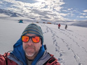 A man in a striped hat an orange sunglasses stands in front of a barren landscape in Antarctica