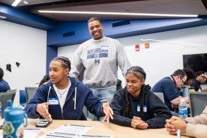 A person wearing a "Michigan IOE" sweatshirt stands and smiles behind two seated individuals at a table in a classroom setting. They appear to be in discussion, with papers and water bottles visible on the table. Other people are engaged in conversation in the background.