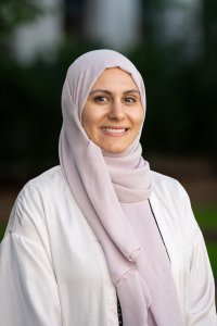 Woman in a hijab smiles at the camera in front of a blurred green background