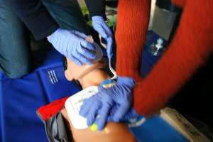 Two hands wearing blue surgical gloves administer CPR on a dummy. CPR training image from Wikimedia Commons.