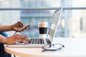Image of person sitting at a short table holding their phone and working on their laptop, with a coffee nearby.
