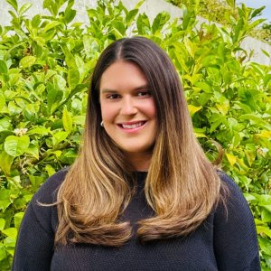 Jenna Birch smiles for a headshot against a background of greenery. She smiles at the camera, wearing a dark colored blouse.