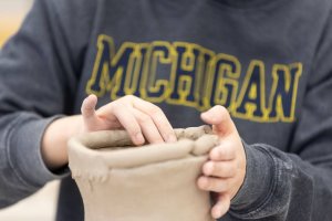 A student wearing a MICHIGAN sweatshirt shapes a clay vessel