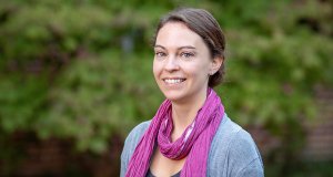 A person is standing outdoors in front of a background of green foliage. The person is wearing a grey cardigan over a dark shirt, accessorized with a bright pink scarf wrapped loosely around their neck. Their hair is pulled back, and they are wearing stud earrings. The setting appears to be calm and natural.