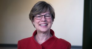 A person wearing a read jacket with a high collar, short hair,  smiling with eye glasses with blue and red frames