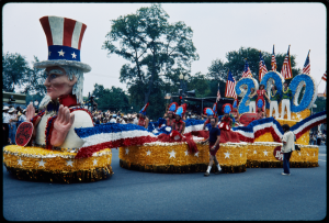 a parade float with a large Uncle Sam in front wearing a stars and stripes top hat and red, white, and blue banners and flags behind him.