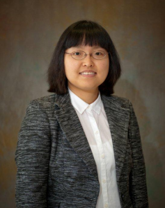 Headshot of person in grey suit with glasses and brown background