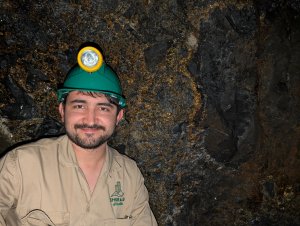 Man in a green hard hat smiles in front of a rock wall with gold deposits running through it