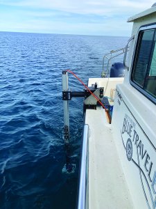 View of a boat on the water. A long, skinny sonar system is mounted vertically to the rear of a vessel, which reads “Survey Vessel Blue Traveler.”
