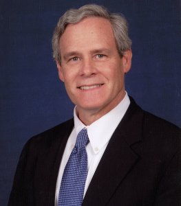 an image of Gleaves Whitney smiling, with wavy grey hair and a black suit and blue tie