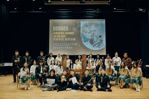 A large group photo of members of the Qingyun Chinese Music Ensemble on a concert stage. Around twenty performers are arranged in several rows, some seated and some standing, holding traditional Chinese musical instruments such as pipa, erhu, guzheng, ruan, and bamboo flutes. A projection screen behind them displays the concert title along with the ensemble’s name. The performers wear coordinated black, white, and green concert attire. The stage has wooden flooring and theatrical lighting, creating a formal concert setting.